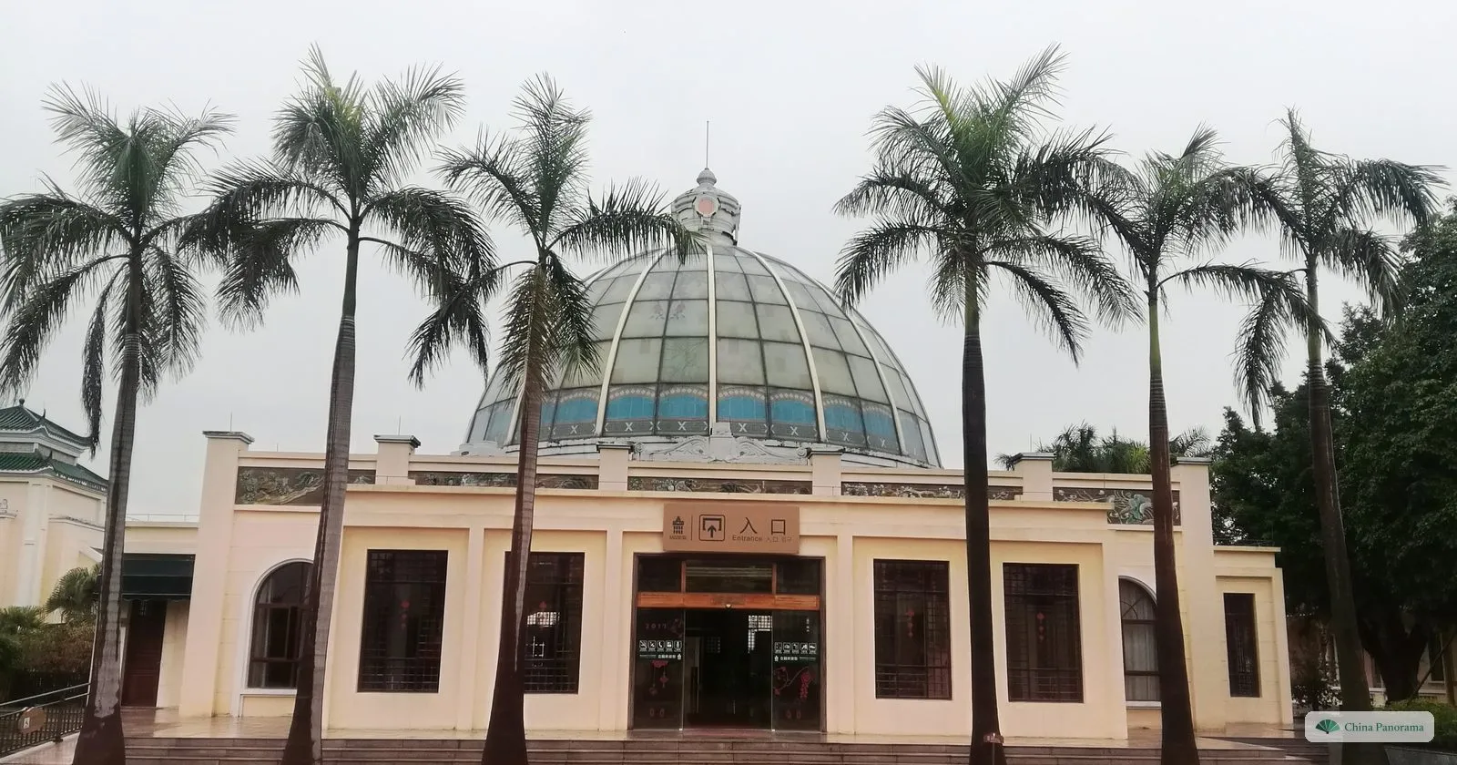 The entrance building of Li Garden in Kaiping, cream-coloured with a large glass dome behind it and tall palm trees in front