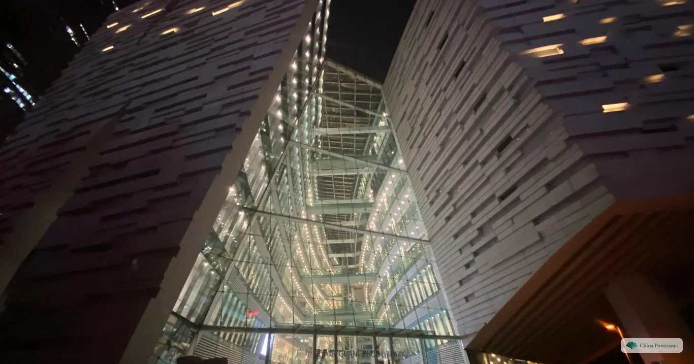 The Guangzhou Library at night from below: two stone-clad blocks leaning apart with a lit glass atrium between them and people at the entrance