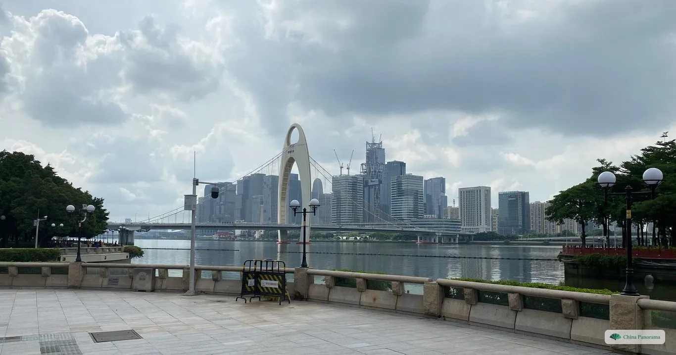 The Liede Bridge across the Pearl River, its single white pylon rising above the deck, with the towers of the city behind and a paved riverside promenade in front