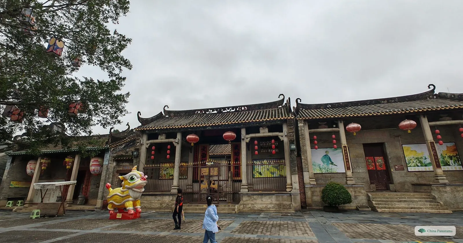Two ancestral halls with upturned roof ridges and red lanterns on a paved square in Lingnan Impression Park, a lion-dance figure by the door