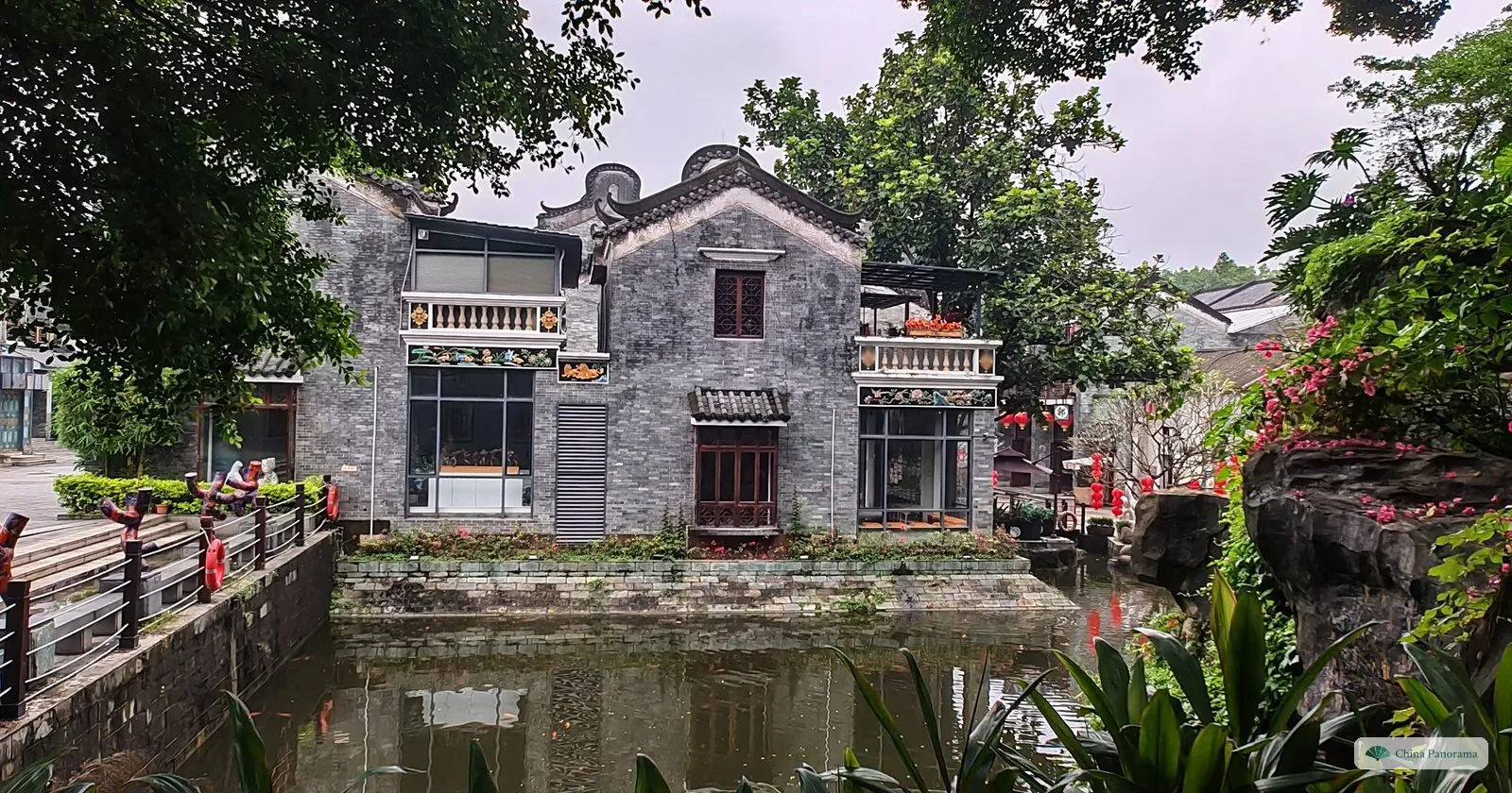 A grey-brick two-storey house with carved balconies beside a pond, with rockery and flowering bushes, in Lingnan Impression Park