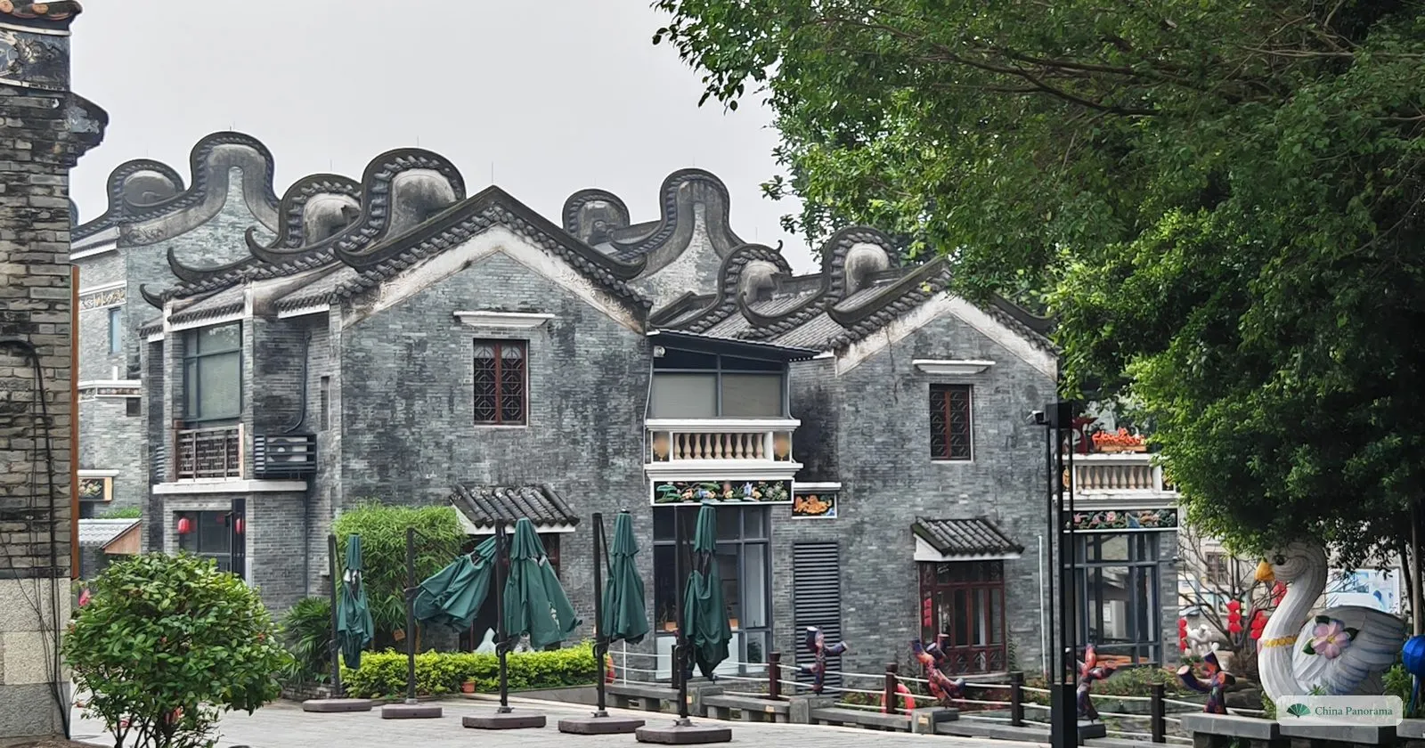 A row of grey-brick houses in Lingnan Impression Park with tall curved gable ends shaped like wok handles, green parasols and a swan sculpture in front