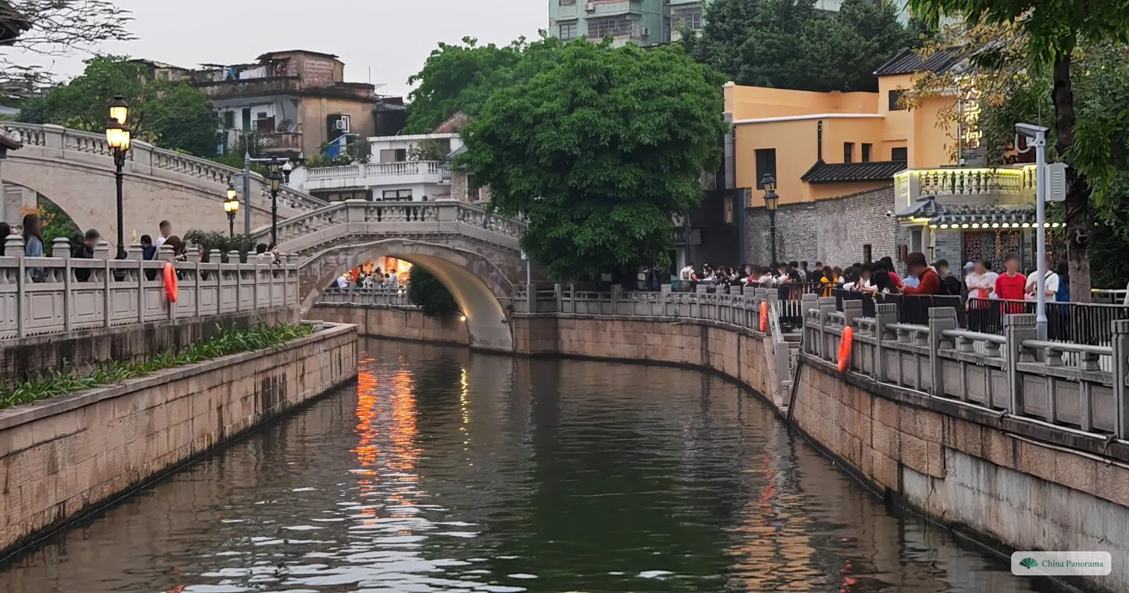 The Lizhiwan canal at dusk with lights reflected in the water, a humpbacked stone bridge in the distance and a crowd along the railing on the right