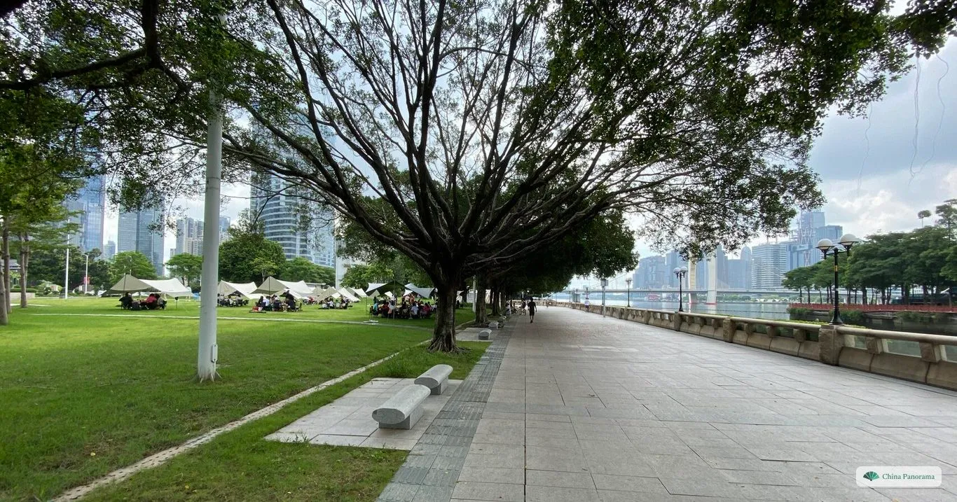 A wide riverside promenade under large banyan trees, with a lawn on one side and the river and distant high-rises on the other