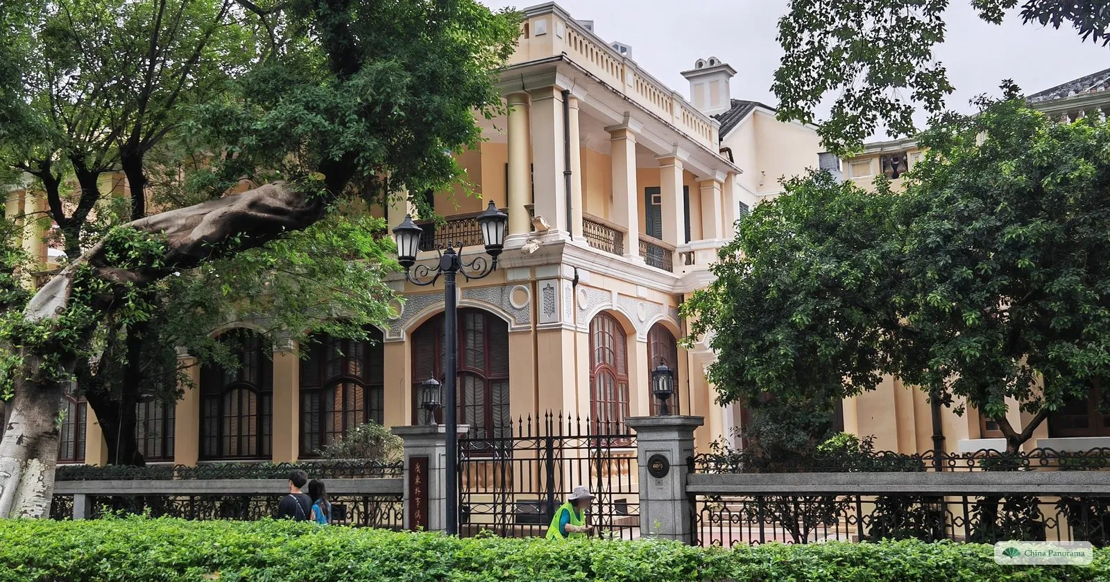 A cream-coloured colonial villa with arched windows and columned verandas behind an iron fence on Shamian Island, half hidden by trees