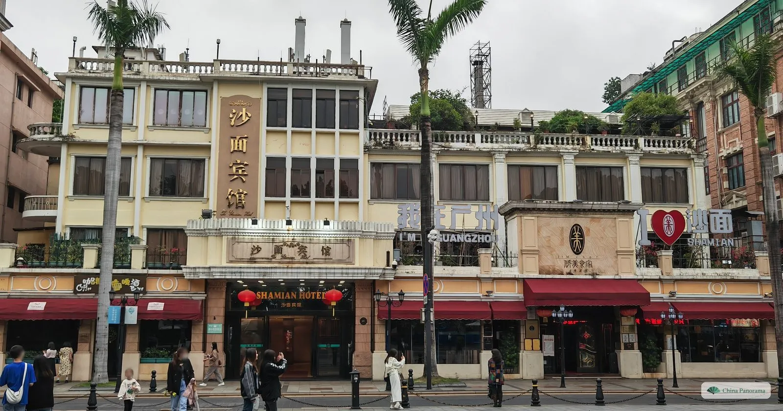 The facade of the Shamian Hotel, a four-storey colonial-era building with balconies and red awnings, palm trees and pedestrians in front