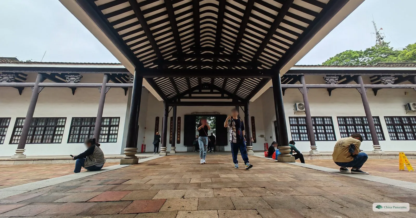 A covered walkway with a dark timber roof between the white two-storey wings of the Whampoa academy, visitors crouching to take photographs