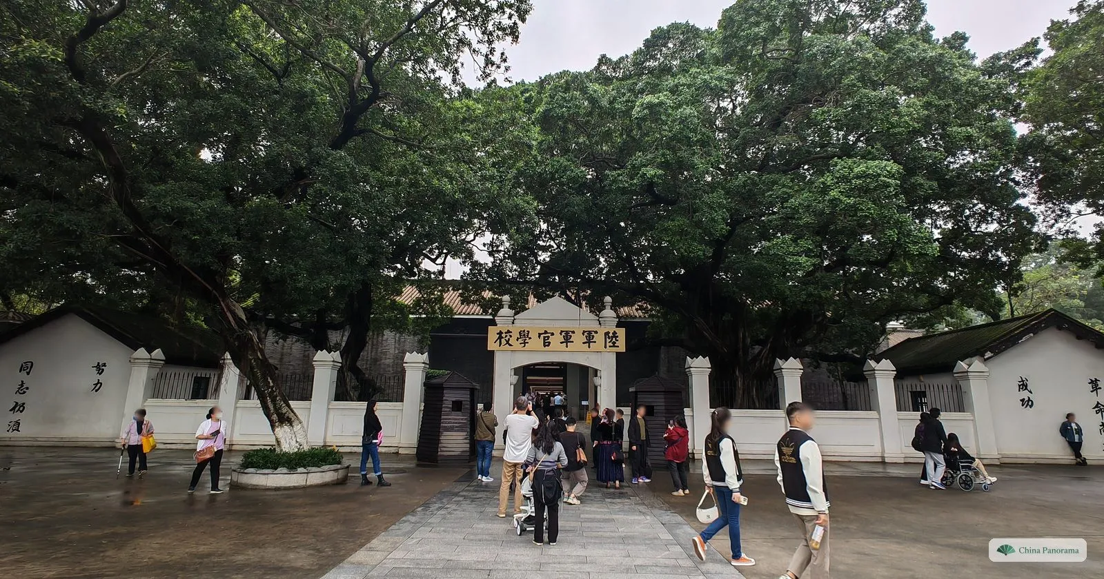 The gate of the Whampoa Military Academy: a white wall with the plaque 陸軍軍官學校 over the entrance, slogans painted on the walls, large trees and visitors walking in