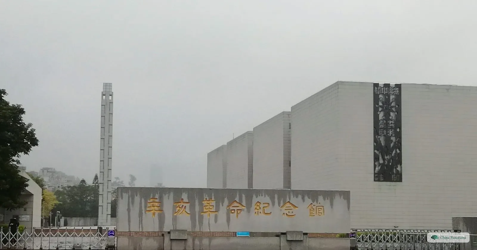 The Xinhai Revolution Museum on a rainy morning: a large pale stone building with the characters 辛亥革命纪念馆 on a low wall and a tall square pillar
