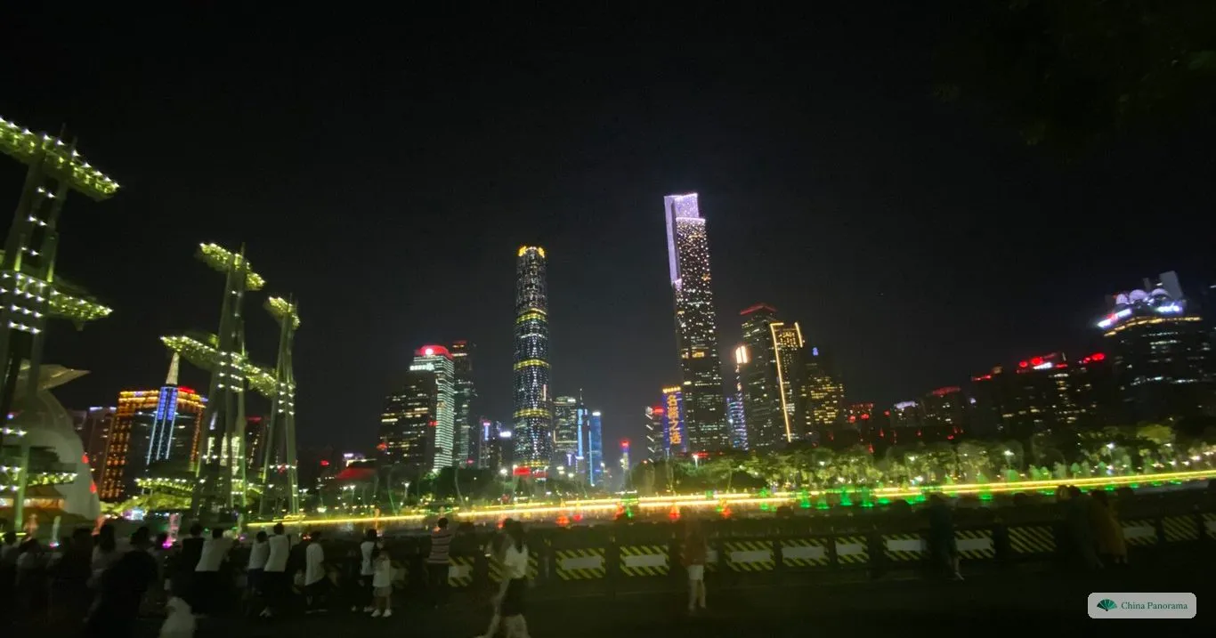 The towers of Zhujiang New Town from Haixinsha at night, with lit masts of the grandstand on the left and people walking along the promenade