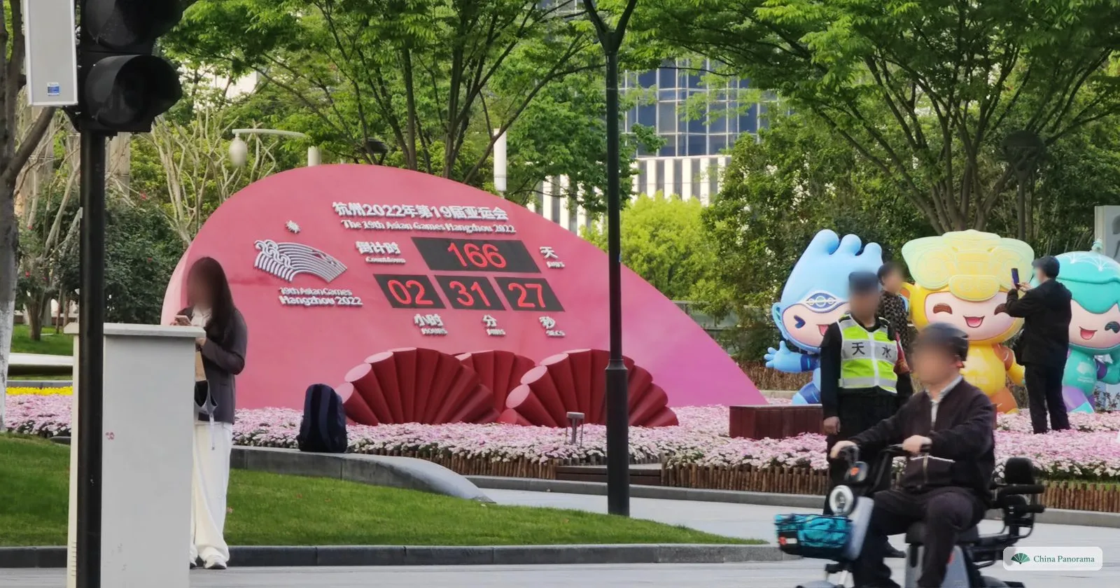 A pink countdown board shaped like a fan showing 166 days, three mascot figures beside it and flowers in front