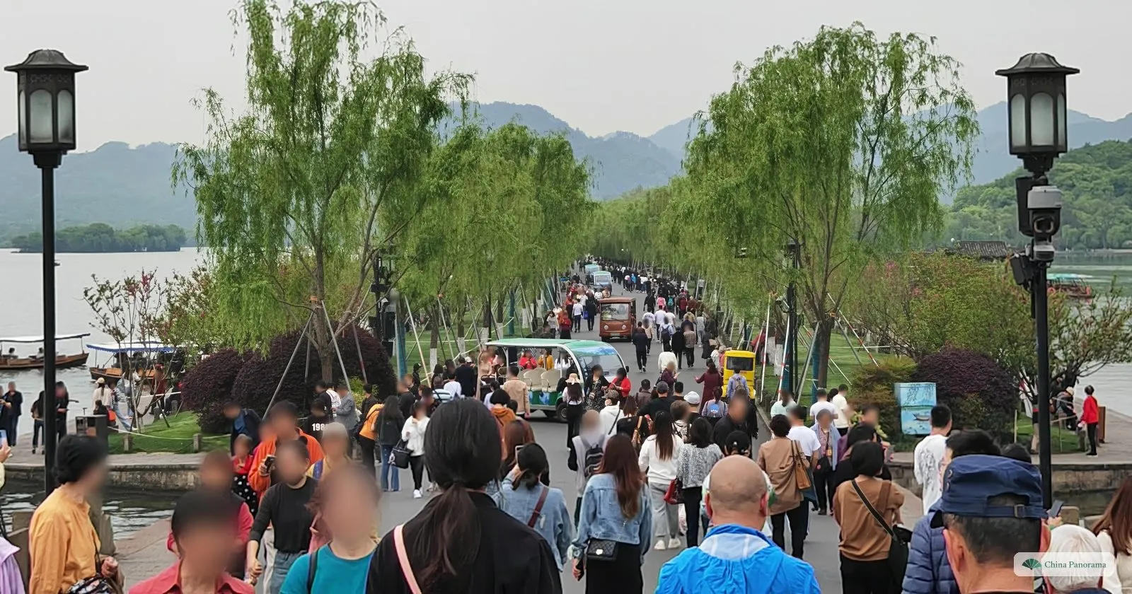 A wide causeway under willows packed with walkers and an electric cart, lake and mountains on either side