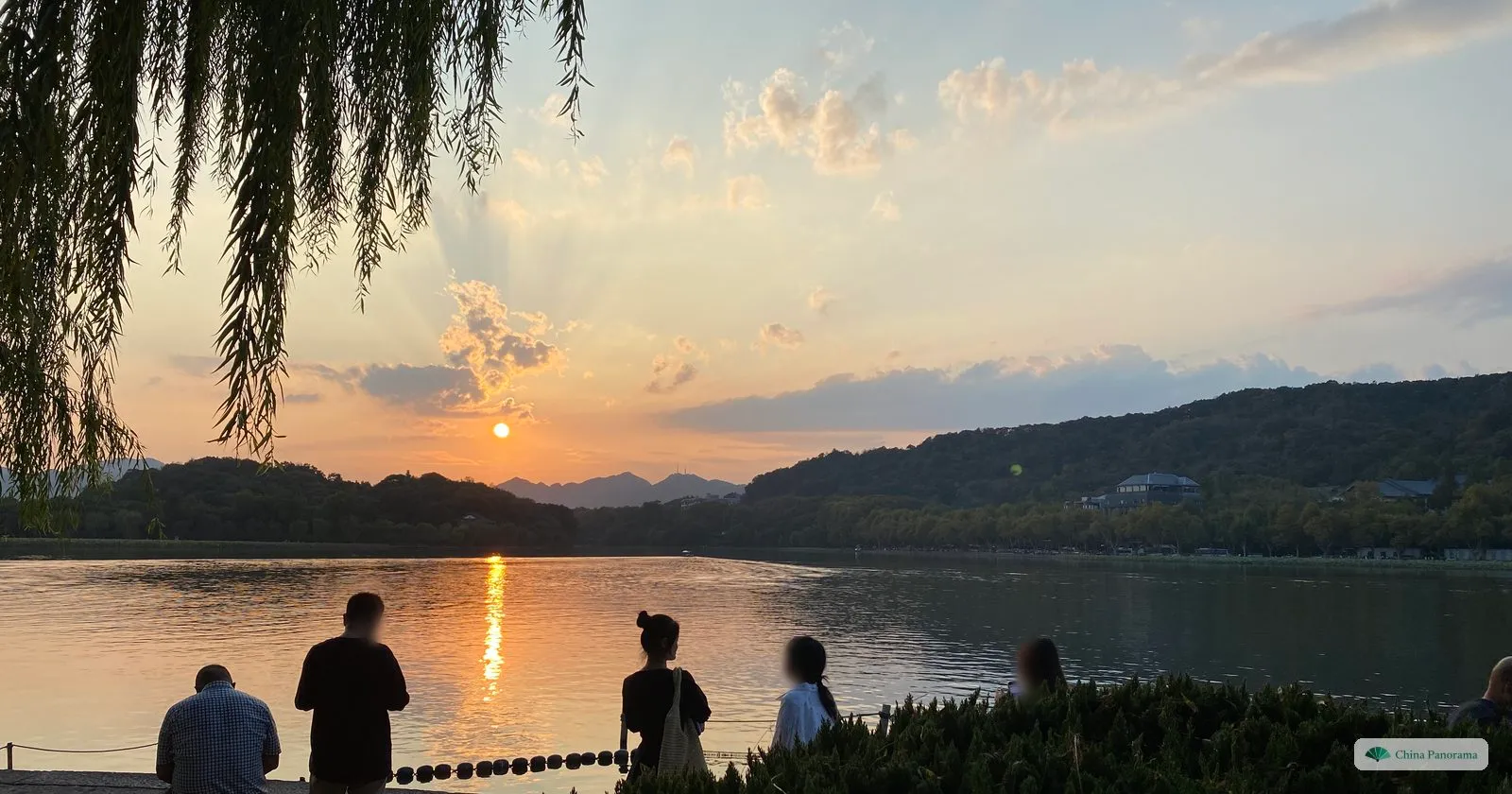 People in silhouette on the Bai Causeway watching the sun set over West Lake behind wooded hills
