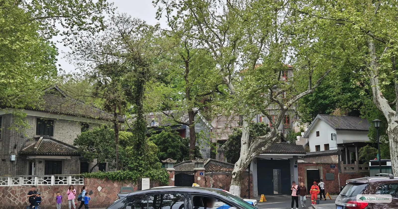 Old brick villas with tiled roofs behind a wall under plane trees, a car passing