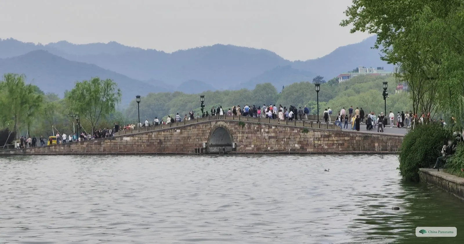 A low stone arch bridge crowded with people, a causeway of willows behind and misty mountains