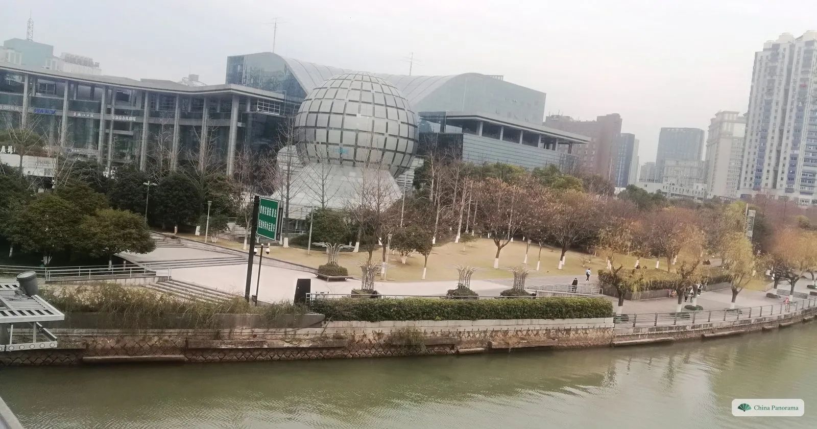A large glass globe on a museum building beside a canal, a square with bare trees and apartment towers behind