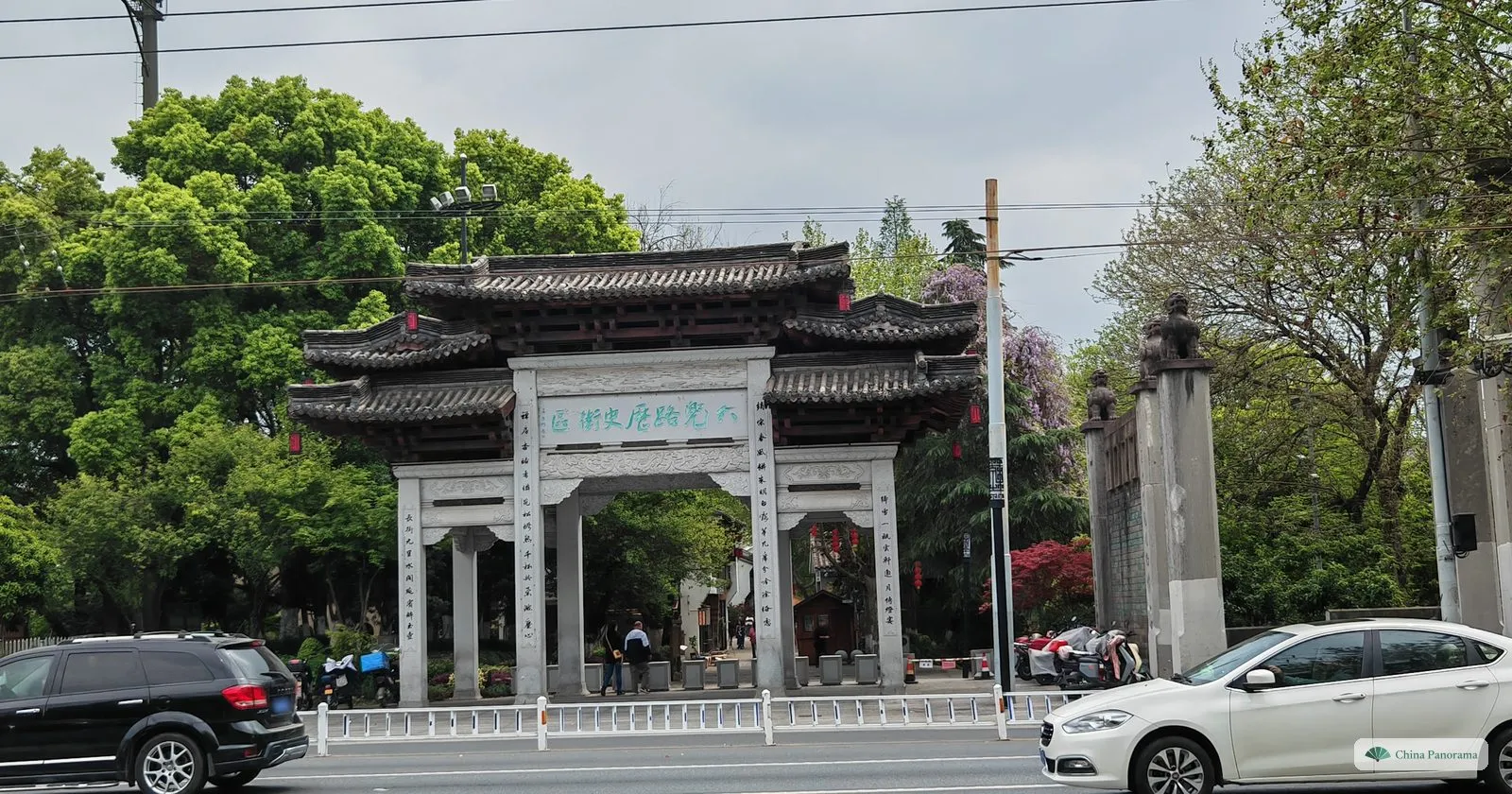 A grey stone memorial archway with tiled roofs and a plaque of green characters beside a busy road