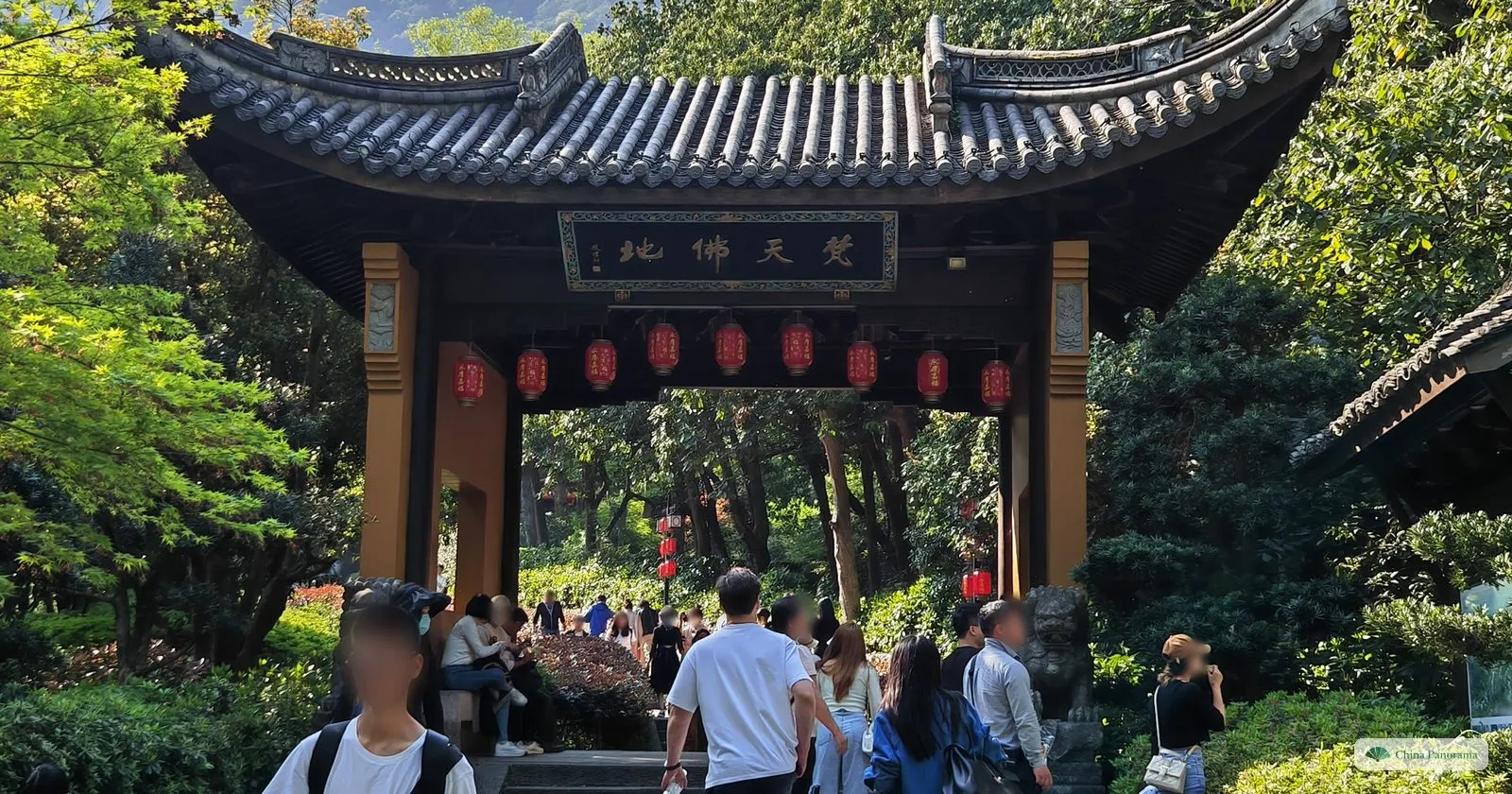 A wooden pavilion gate with a plaque of four characters and a row of red lanterns, visitors walking through to a garden