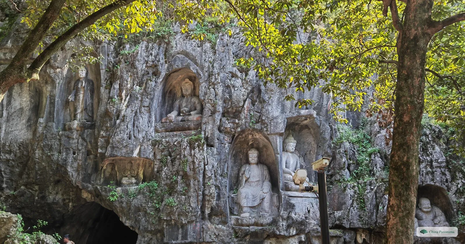 A grey limestone cliff with Buddhist figures carved in niches, a cave mouth at the bottom left