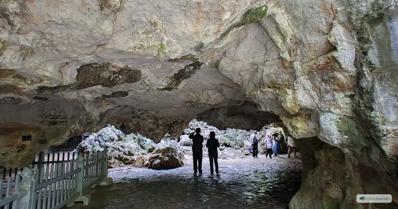 Inside a limestone cave with a low rock ceiling, small carved figures on the wall and people silhouetted against the light at the far end