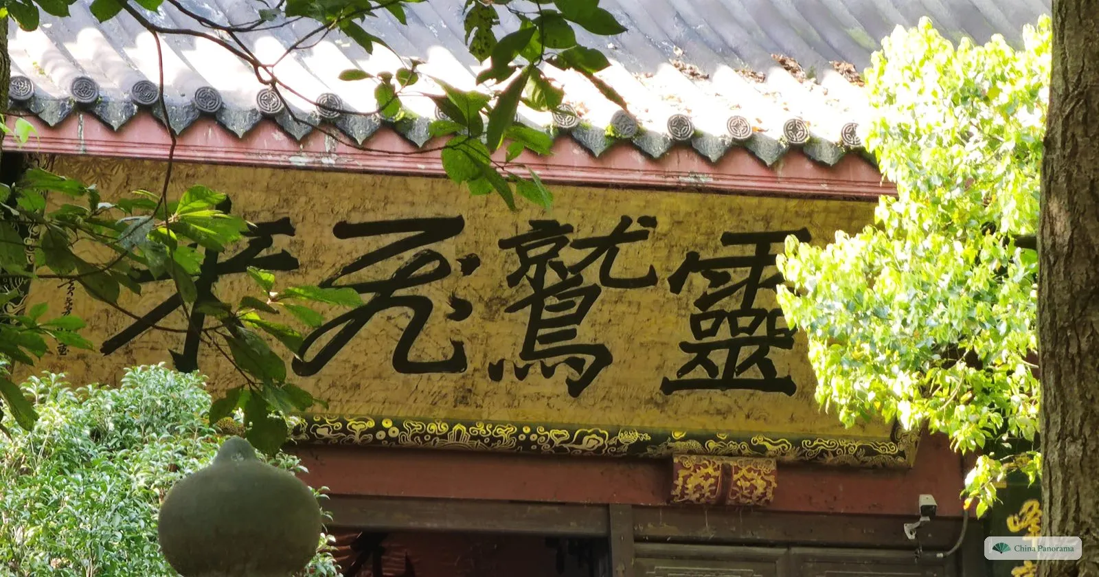 A carved stone panel of four large dark characters on yellow under the eaves of a roof, leaves in front