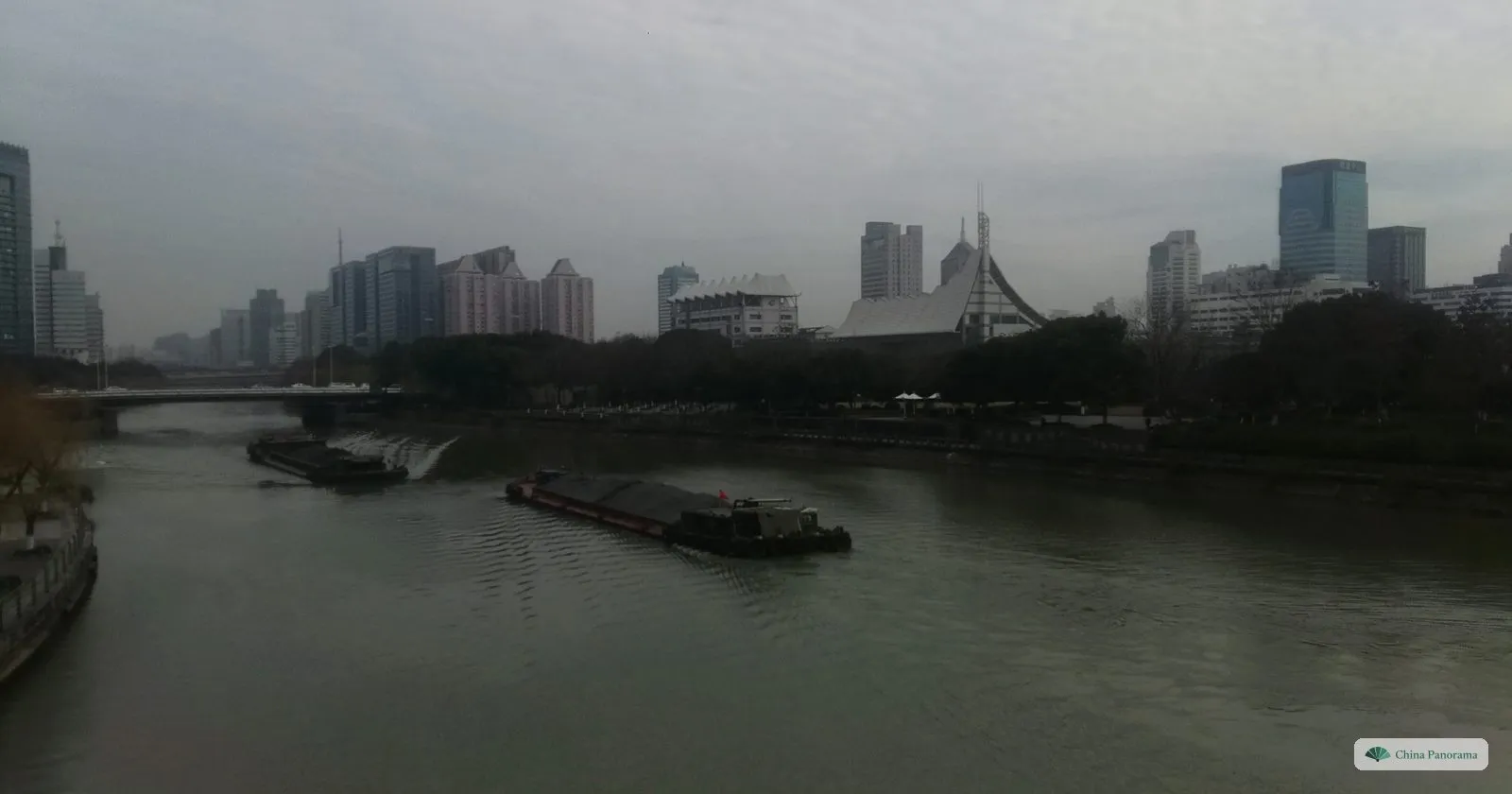 Two long low cargo barges on a wide grey waterway between city buildings on a hazy day