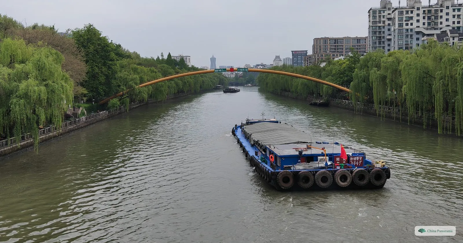 Looking along a straight waterway lined with willows, a loaded barge in the middle and a yellow arched bridge in the distance