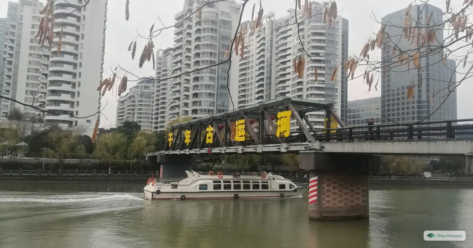 A white passenger boat speeding under a steel bridge hung with large yellow characters, apartment towers behind