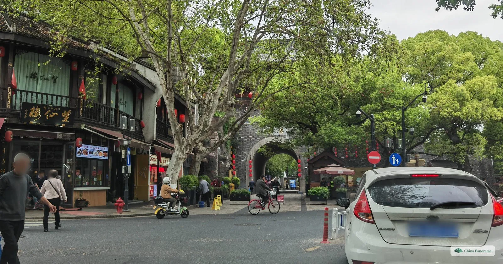 A street of old two-storey wooden houses leading to a stone city gate with an arch, a white car in the foreground