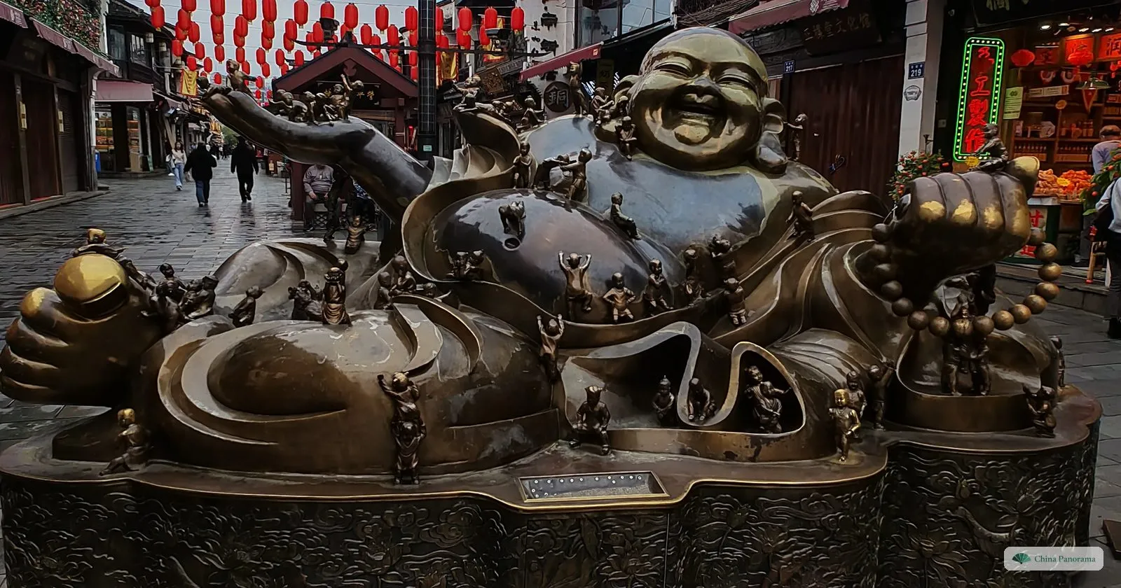 A large bronze statue of a reclining laughing Buddha covered with small climbing children, on a pedestrian street