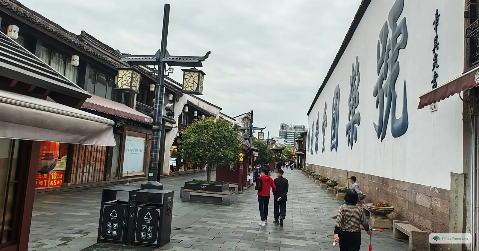 A very long white wall along a pedestrian lane, painted with enormous dark characters, people walking past