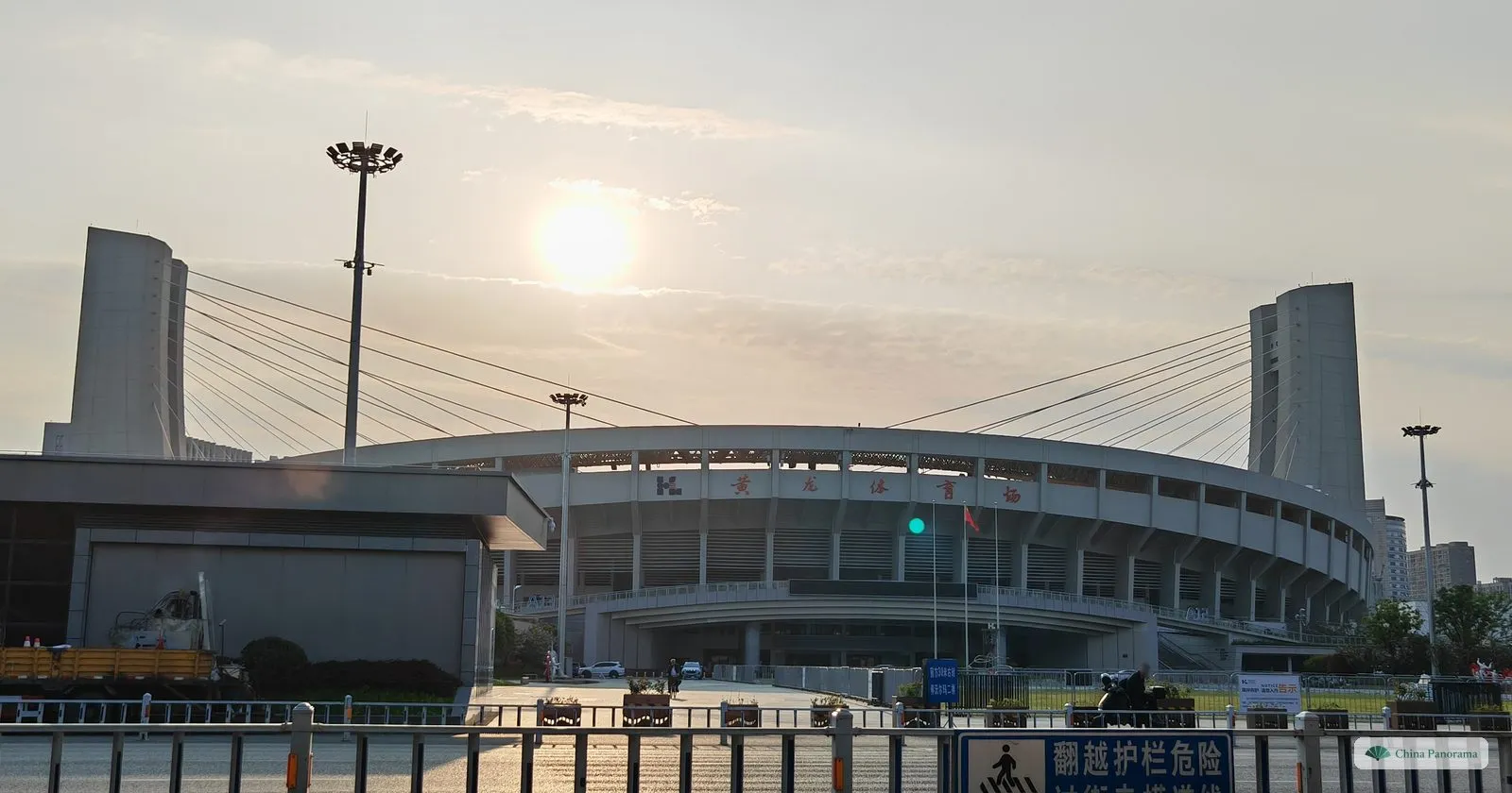 A large stadium hung by cables from two tall concrete pylons, the sun low in a hazy sky