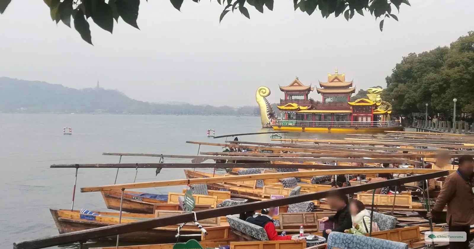 Wooden rowing boats moored in rows on a grey lake, a large ornate pleasure boat with golden roofs behind and a hill with a thin pagoda in the haze
