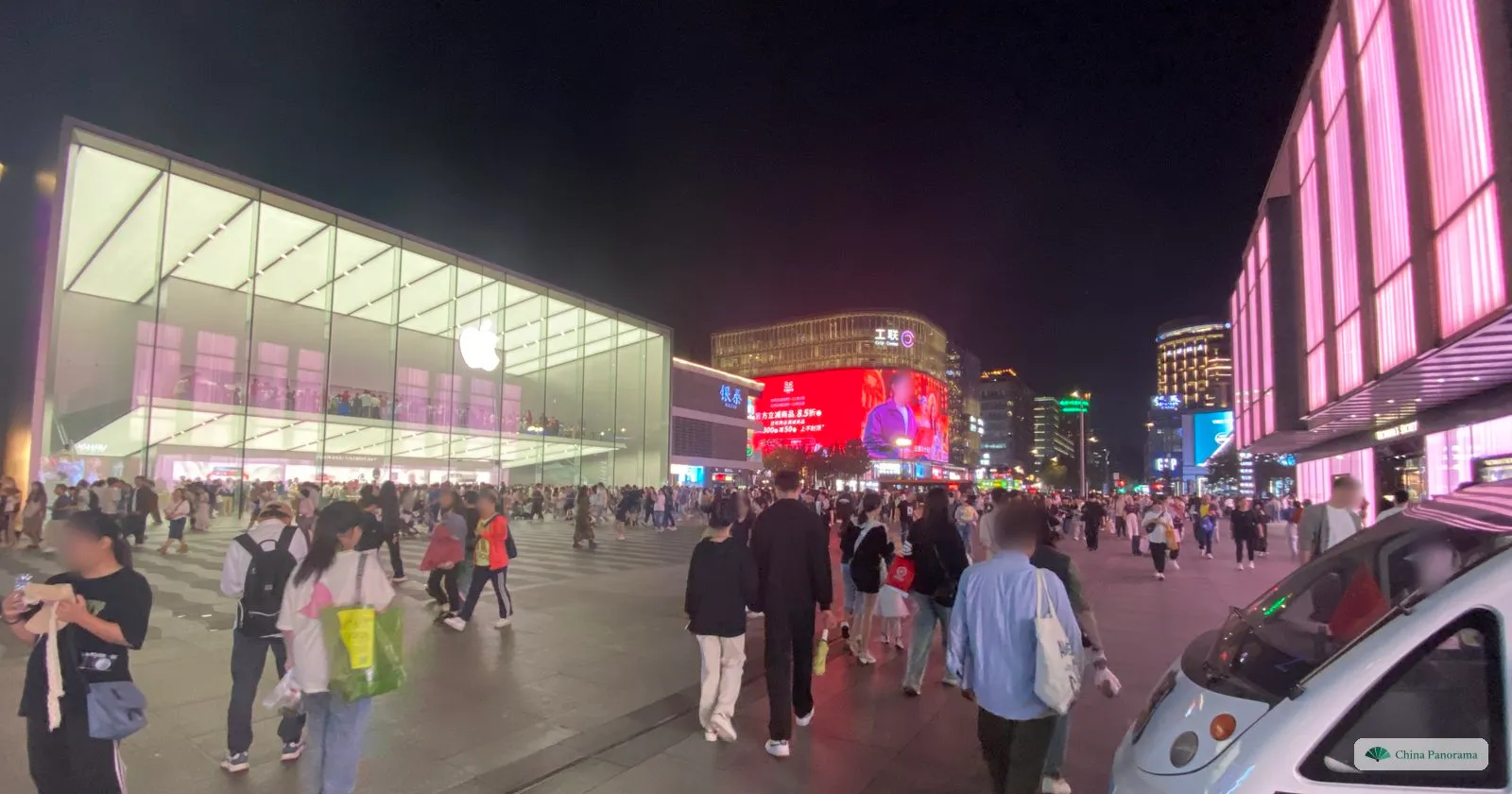 A crowded pedestrian square at night beside a large glass-fronted shop, bright screens on the buildings