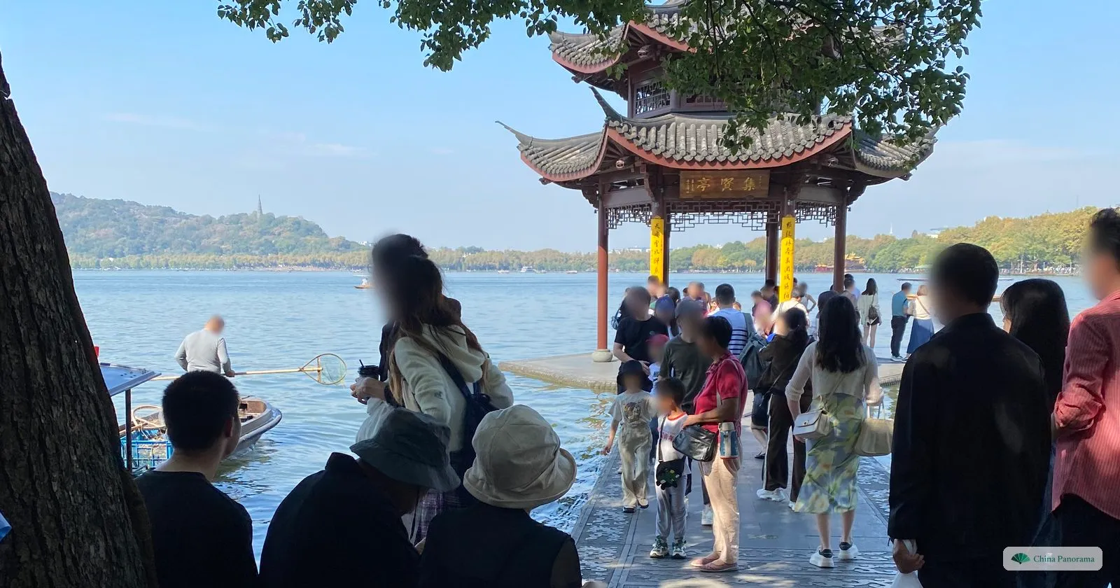 A two-tiered pavilion on a stone platform in a lake, people on the promenade and a boatman with a net, a hill with a pagoda across the water