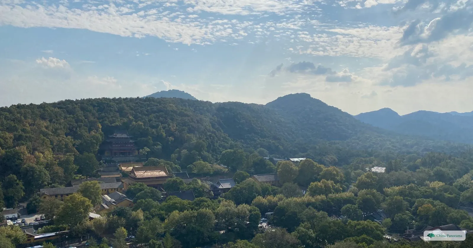 Looking down from a height over wooded hills and the yellow-walled halls of a temple, mountains behind