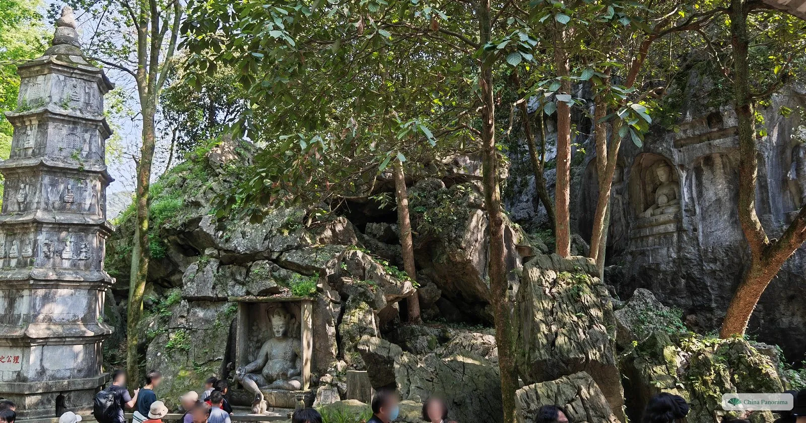 A weathered six-sided stone pagoda carved with small figures beside a rock face with a carved seated figure in a niche, visitors below