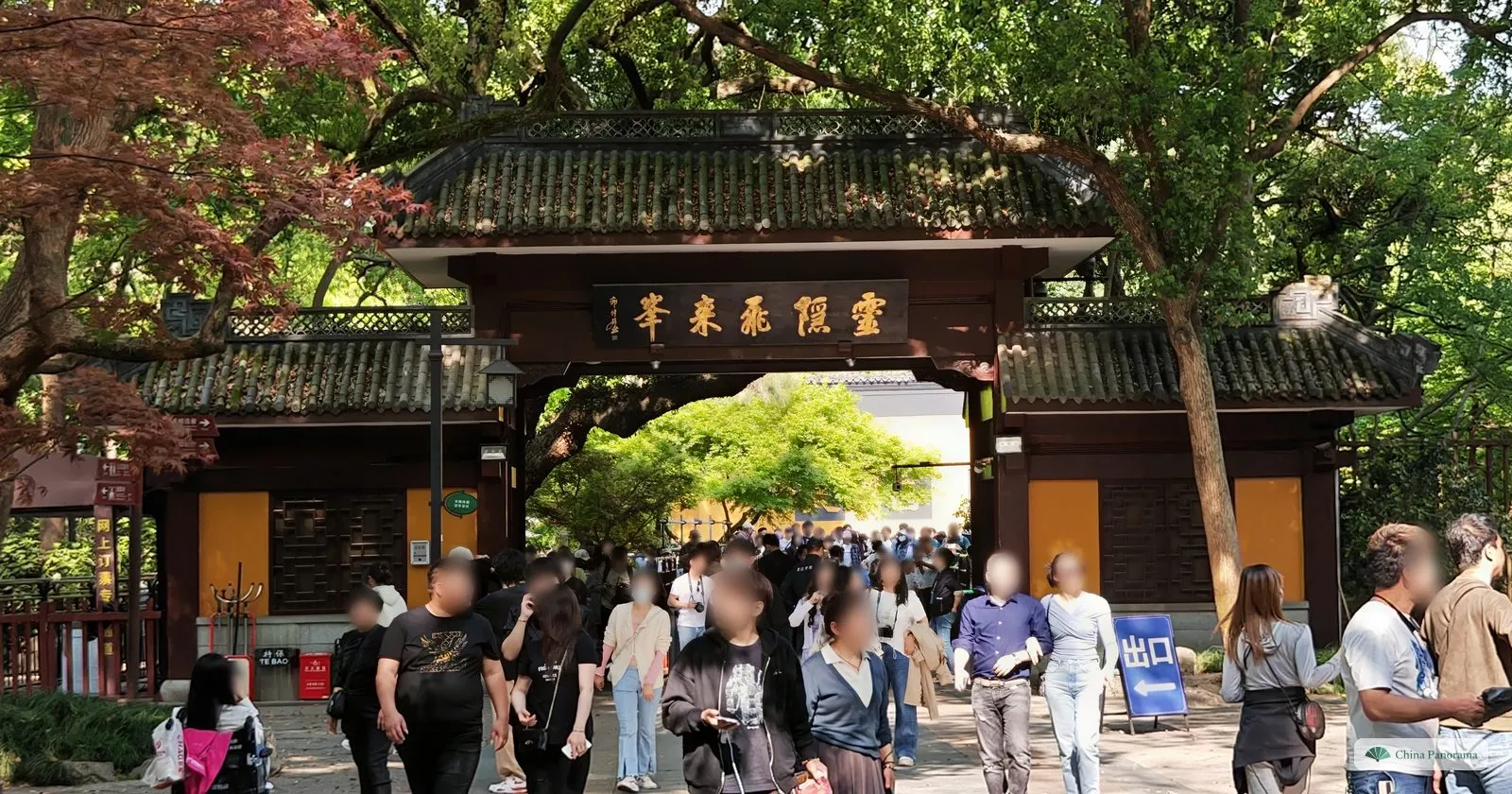 A dark wooden gate building with a black plaque of gold characters, a crowd walking out under trees