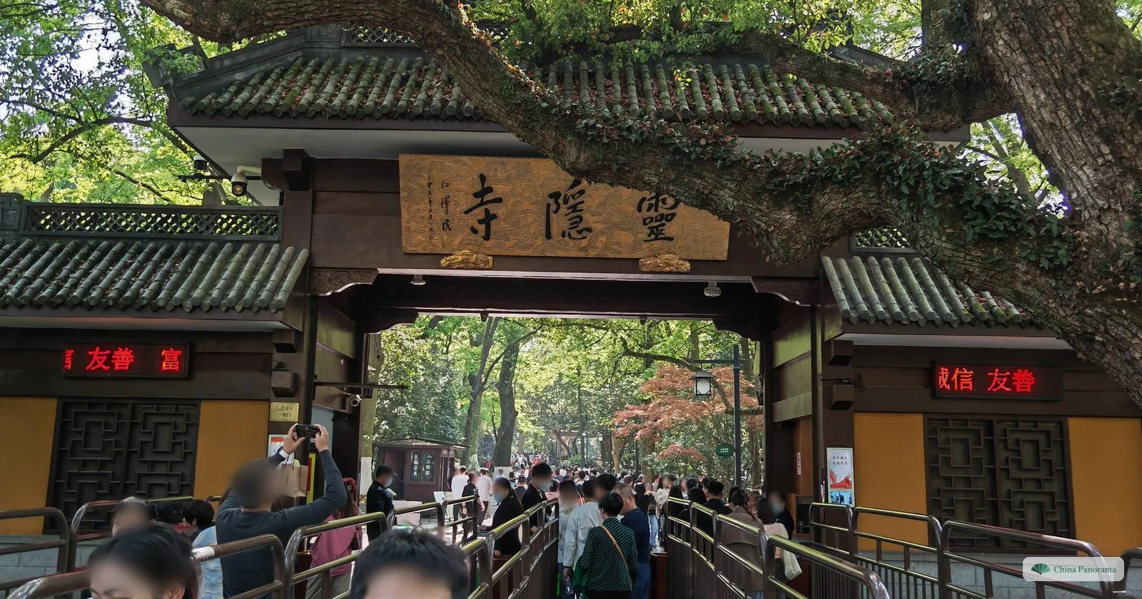 A wooden gateway with a large horizontal plaque of three gold characters, crowds queueing through railings under an old tree