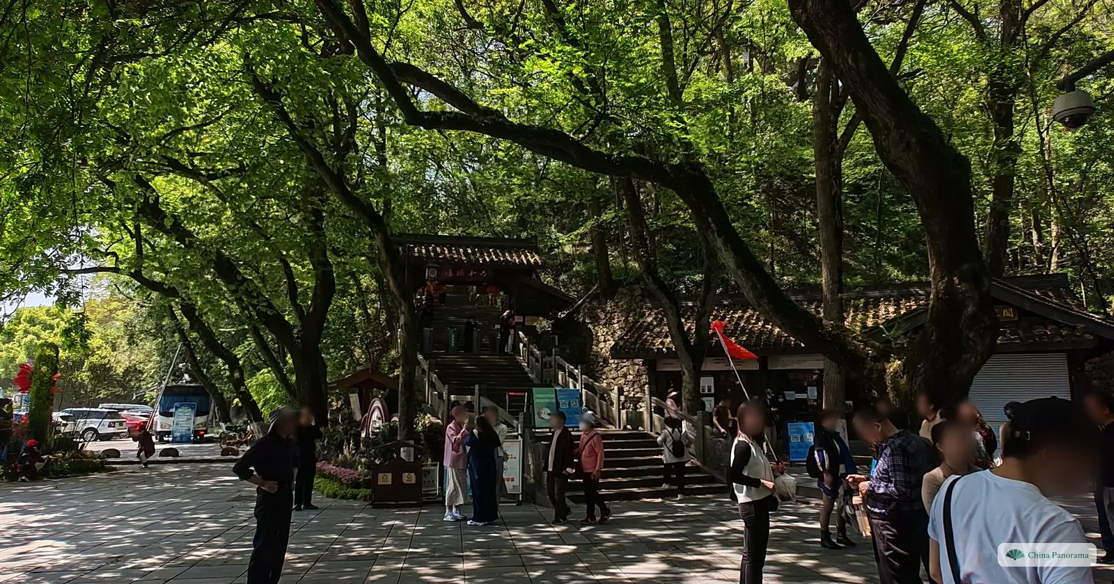 A shady square under spreading old trees with visitors, steps up to a gate building and a red flag