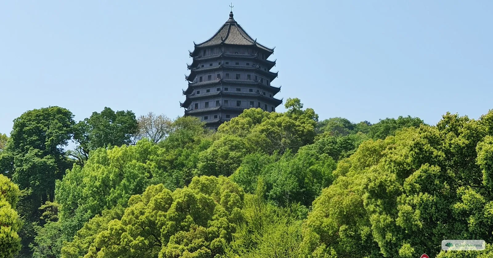 The upper storeys of a tall octagonal pagoda with many tiers of dark eaves rising above a green wooded hill under a blue sky