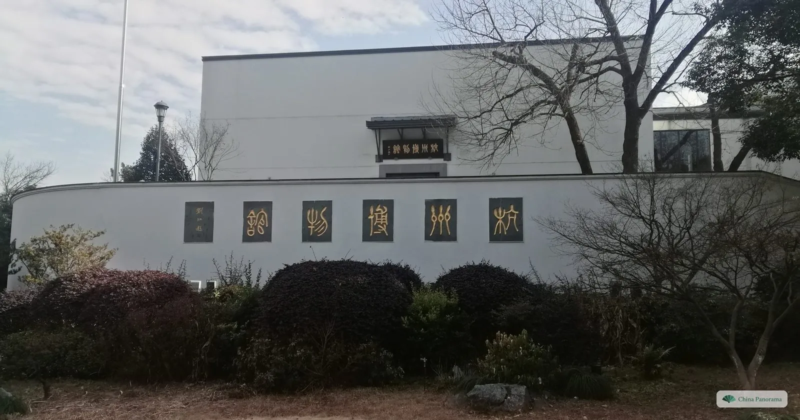 A long white wall with a row of dark square panels of gold characters, a modern white building and bare trees behind