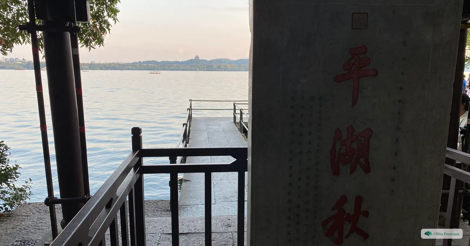 A stone pillar carved with four red characters on a lakeside terrace, a jetty and the lake beyond in evening light