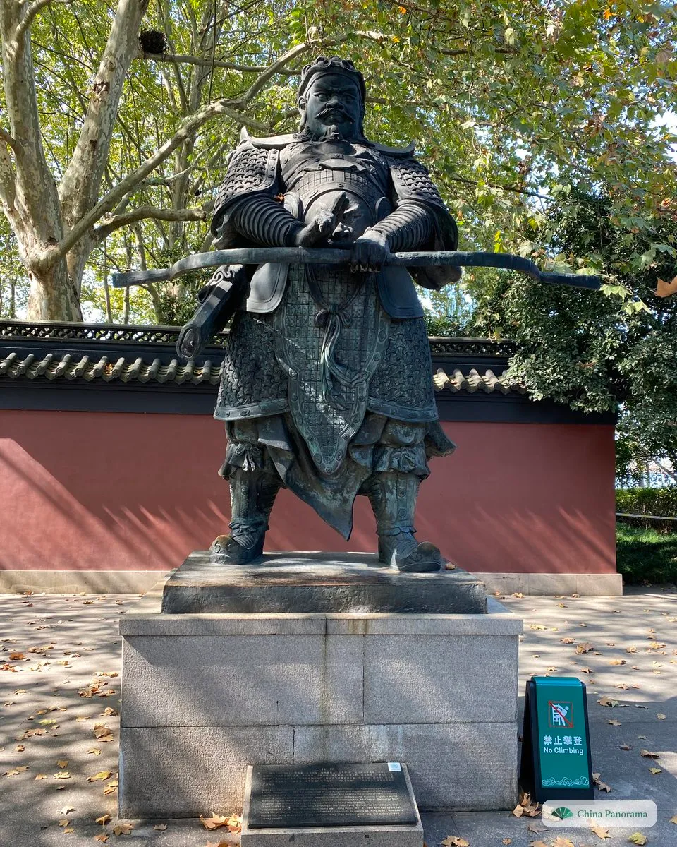 A bronze statue of a warrior in armour holding a bow across his body, on a stone plinth in front of a red wall