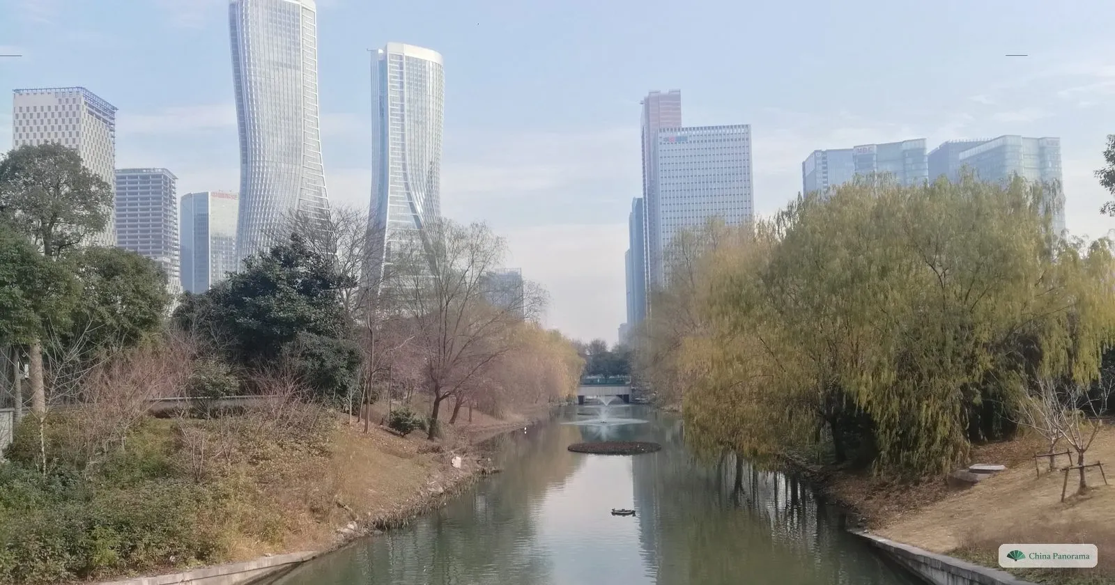 A narrow canal between willows and bare winter trees, tall glass towers of a business district behind