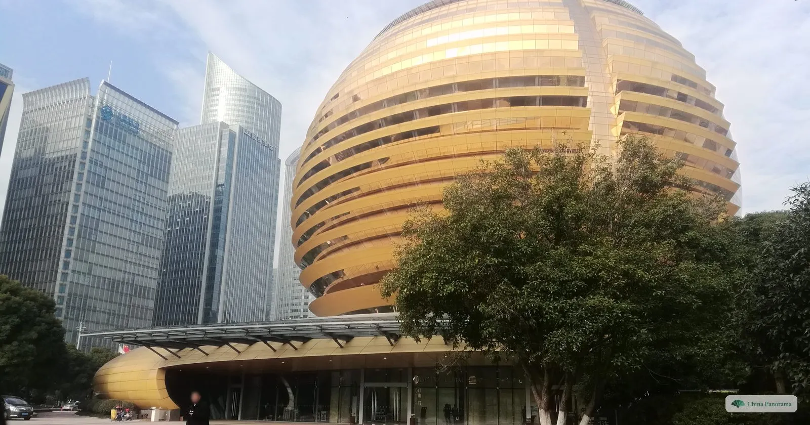 A huge golden sphere of a building with horizontal bands of windows beside glass office towers, a tree in front
