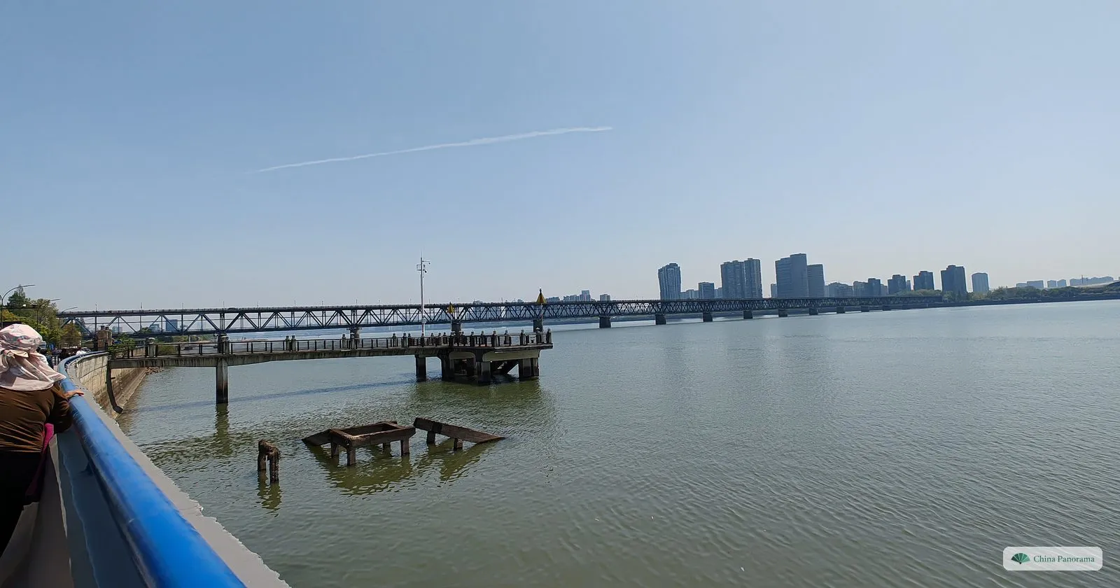 A long steel truss bridge with two decks across a wide calm river, a pier and old concrete posts in the water, high-rise buildings on the far bank