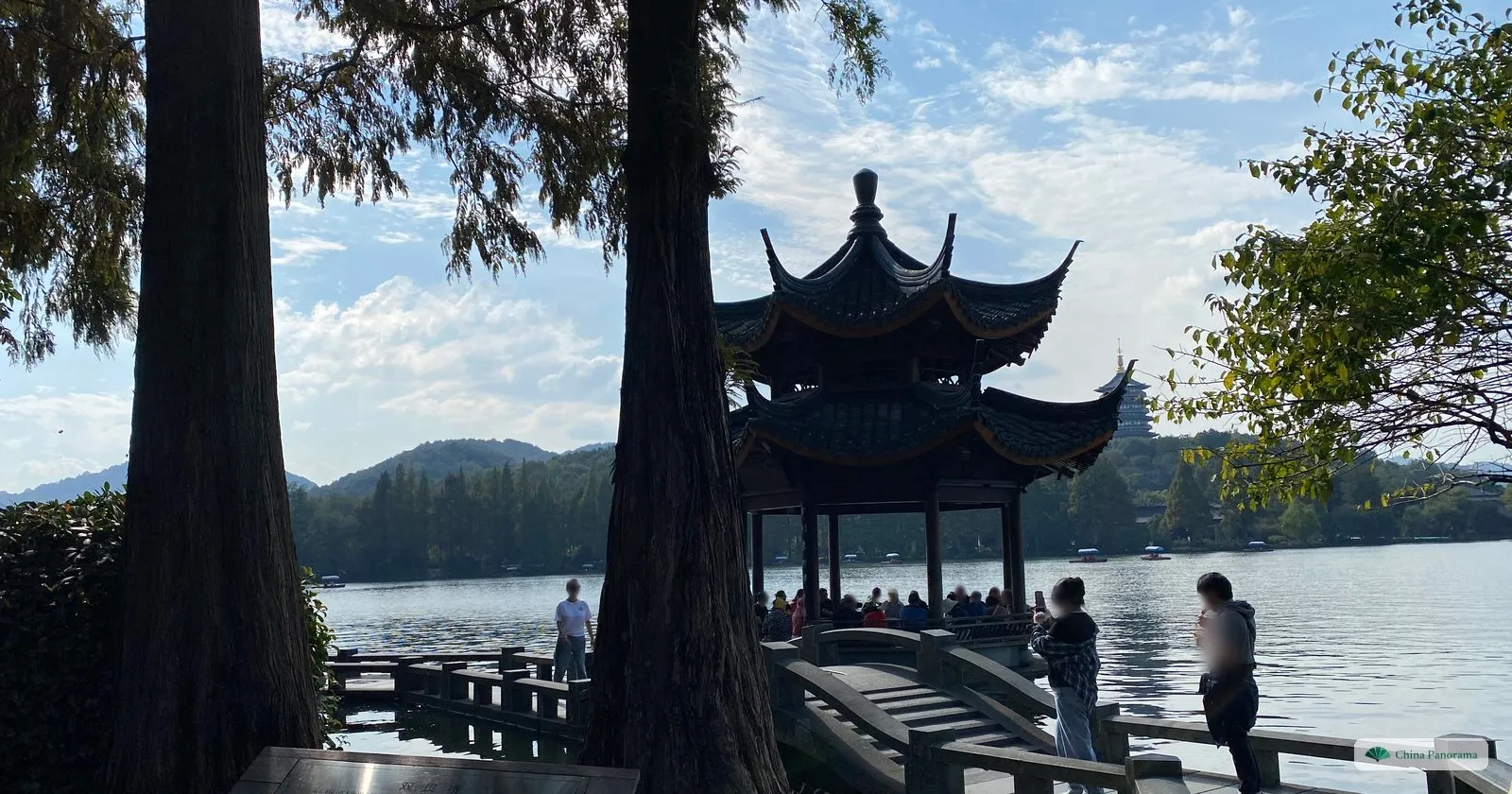 Two tall tree trunks framing a hexagonal two-tiered pavilion on the water, reached by a little arched stone bridge, a tall pagoda in the distance