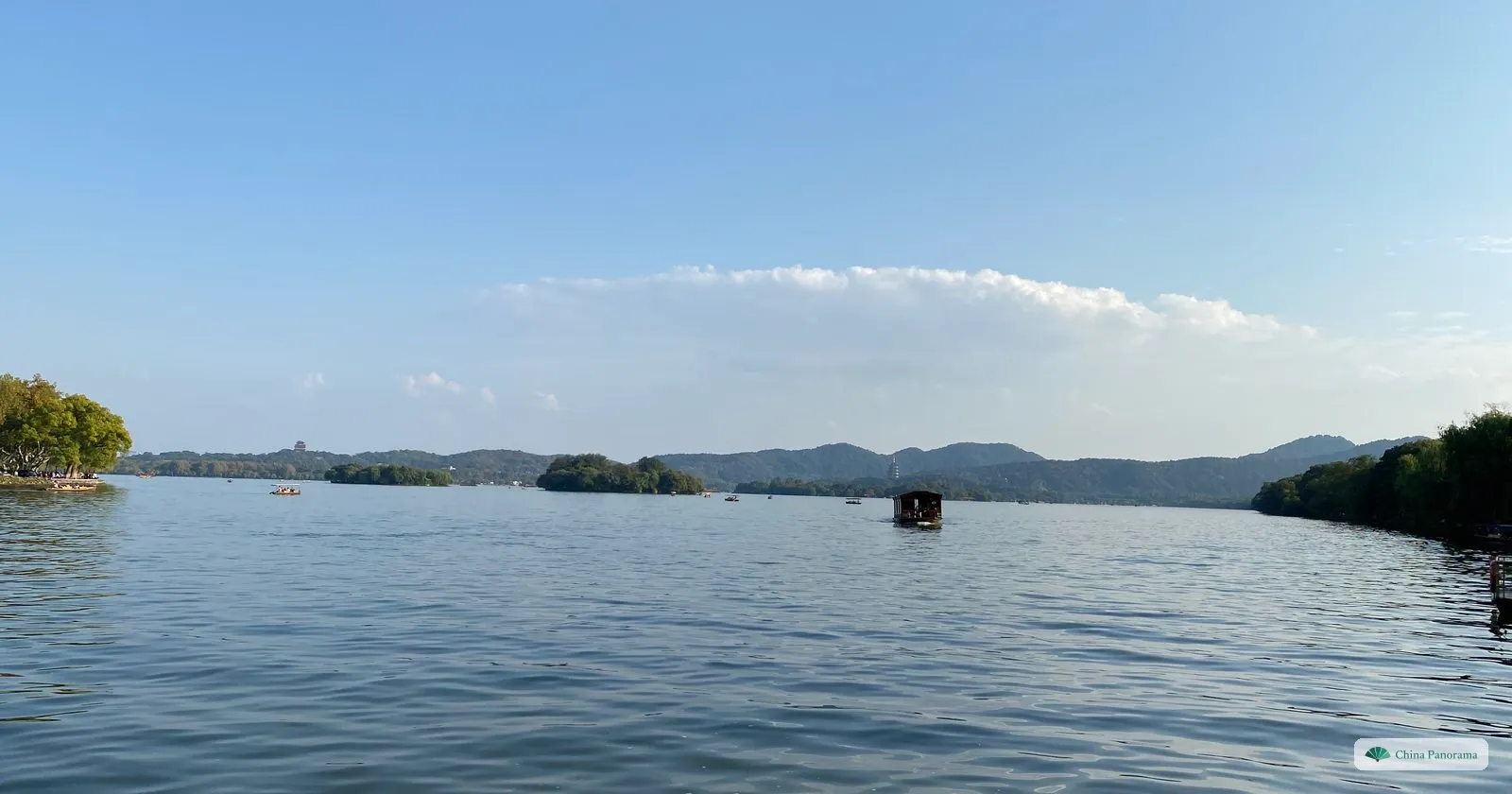 A wide calm lake with a few small islands and a low line of hills under a pale blue sky
