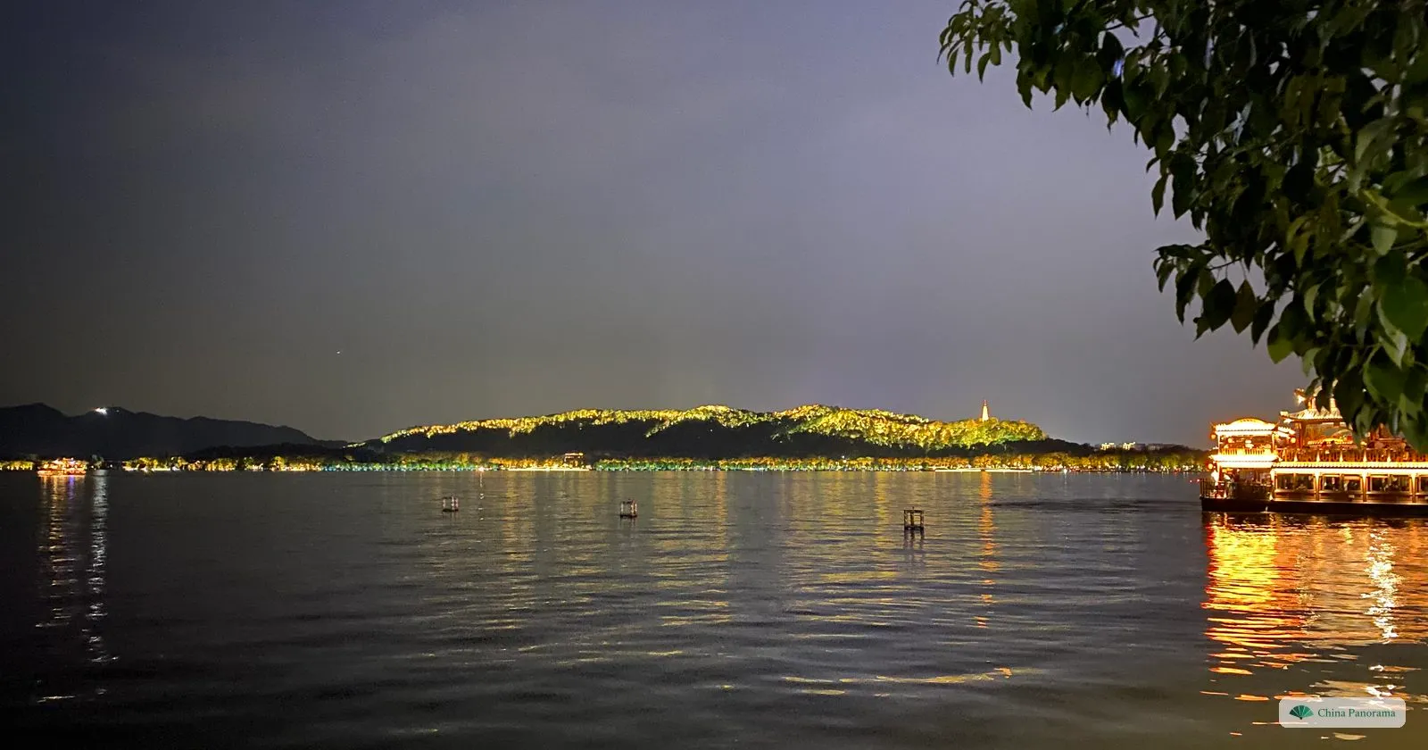 A dark lake at night with a wooded hill lit up along its slopes and a lit pagoda on the ridge